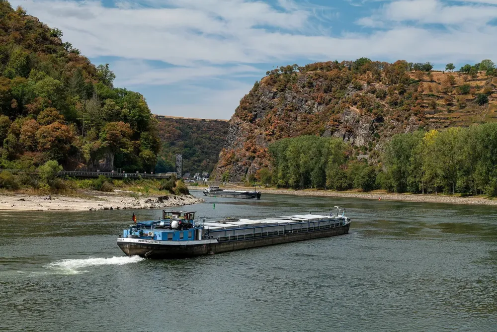 Frachtschiff fährt auf einem Fluss vorbei an felsigen Ufern mit Bäumen.