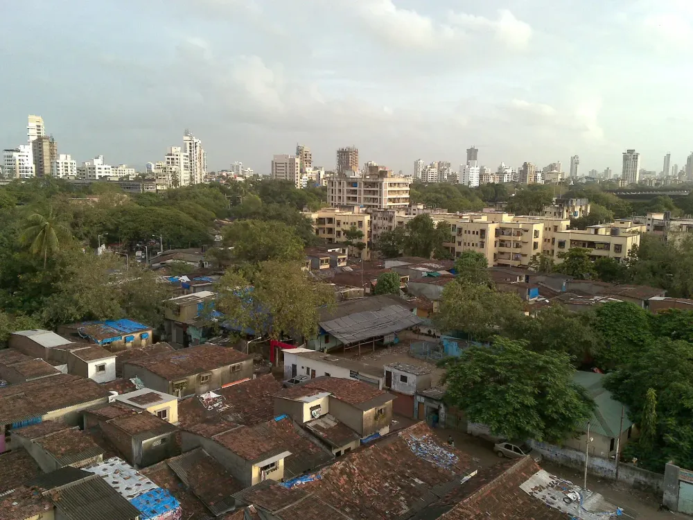 Aerial view of a dense urban slum with brown roofs and distant high-rise buildings.