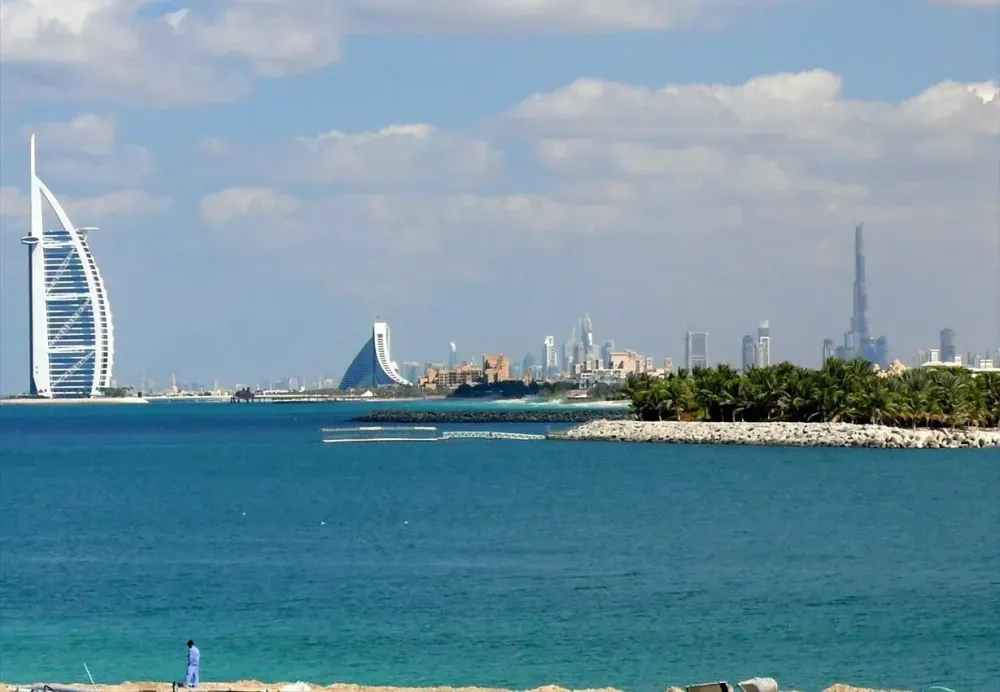 Distant view of the Dubai skyline from the water, featuring the Burj Al Arab and the Burj Khalifa under a blue sky.