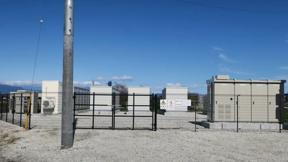 Umzäunte Freifläche mit mehreren weißen Batteriespeicher-Containern, Metallzaun, Strommast und Warnschildern unter blauem Himmel.