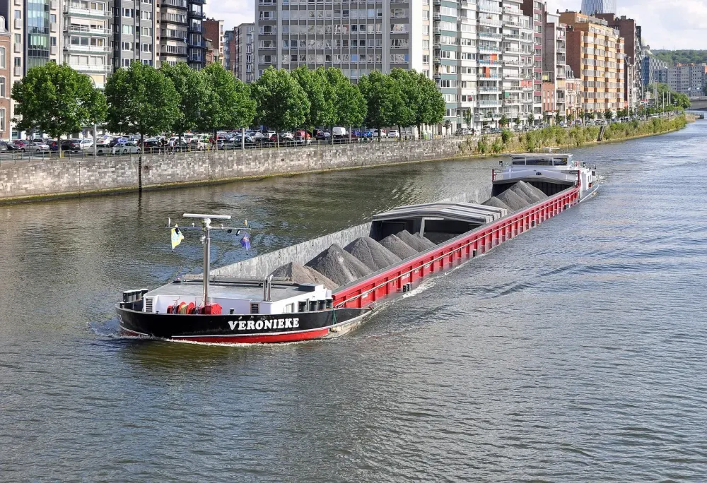 Ein langes Frachtschiff mit Kies auf einem Fluss vor einer Uferpromenade.