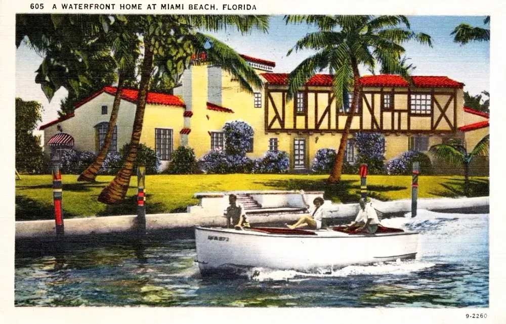 A vintage postcard of a yellow two-story house on a waterfront in Miami Beach with palm trees and a passing motorboat.