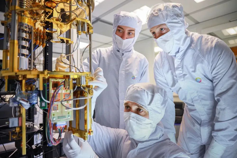 Three people in white cleanroom suits and masks inspecting a gold-colored quantum computing apparatus with wiring in a laboratory.