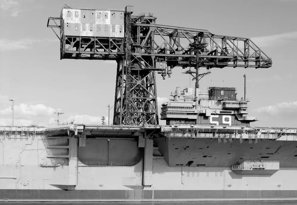 Schwarz-Weiß-Foto eines riesigen Werftkrans über einem US-Flugzeugträger in einem Hafen.