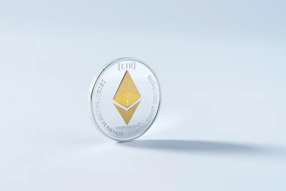 A silver physical coin with a gold Ethereum logo stands on its edge on a light blue surface.
