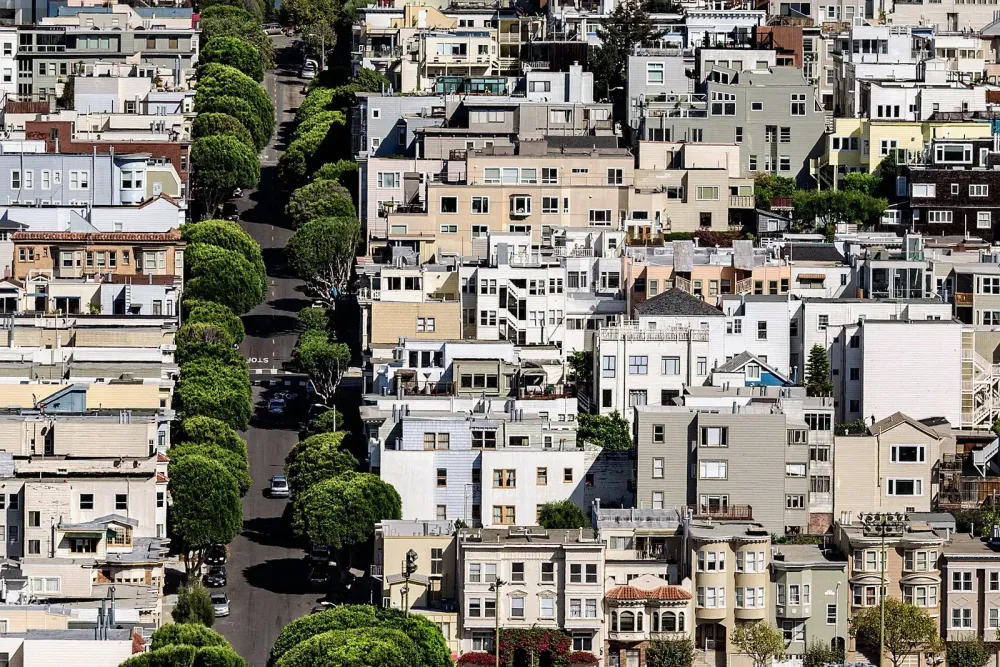 Dense residential buildings along a steep street lined with green, rounded trees in an urban setting.