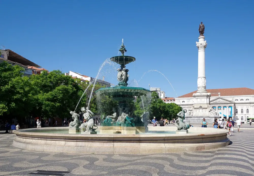 Bronze tiered fountain in a Lisbon square with wave-patterned pavement, a column monument, and yellow classical buildings.