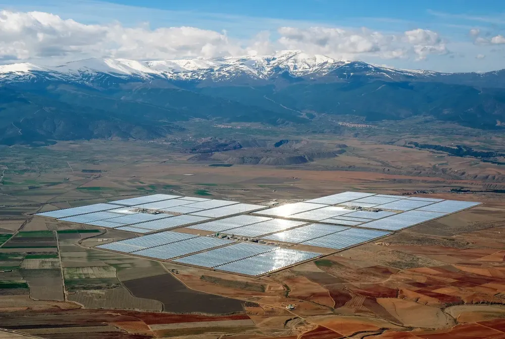Luftaufnahme eines weitläufigen Solarkraftwerks mit spiegelnden Paneelen vor einer Kulisse aus schneebedeckten Bergen.