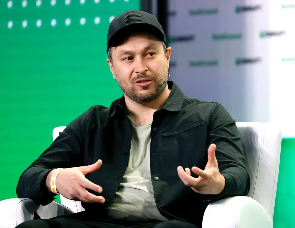A man in a black cap and shirt gestures with his hands while sitting in a white armchair at a conference.