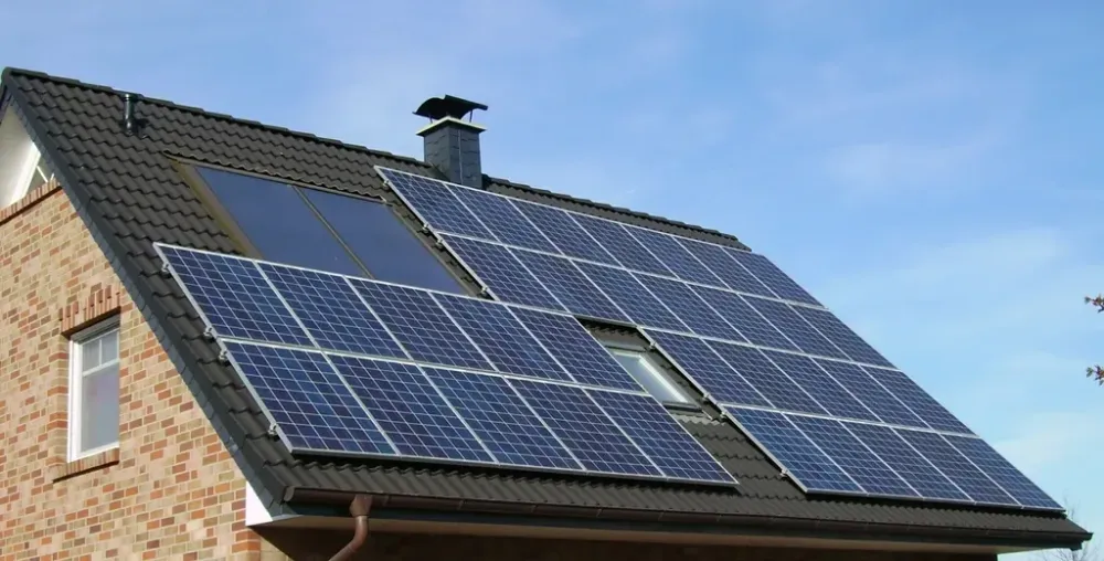 Schräges Ziegeldach eines Backsteinhauses mit montierten Solarmodulen, Kamin und Dachfenstern vor blauem Himmel.