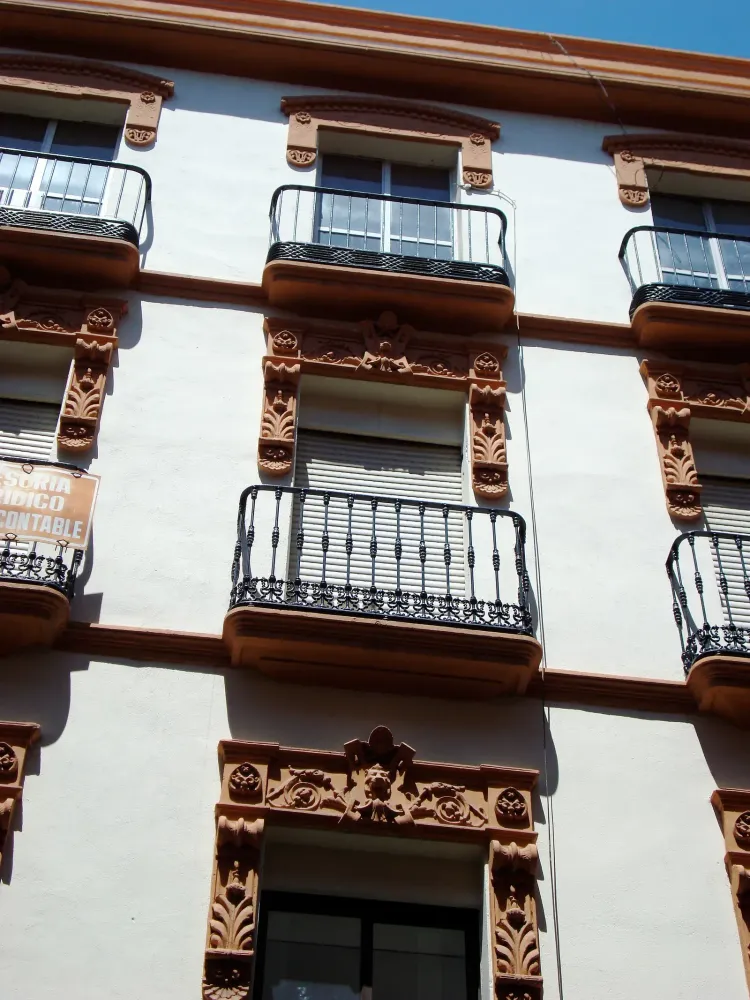 Facade of a white Spanish apartment building with ornate terracotta trim, wrought-iron balconies, and a partial street sign.