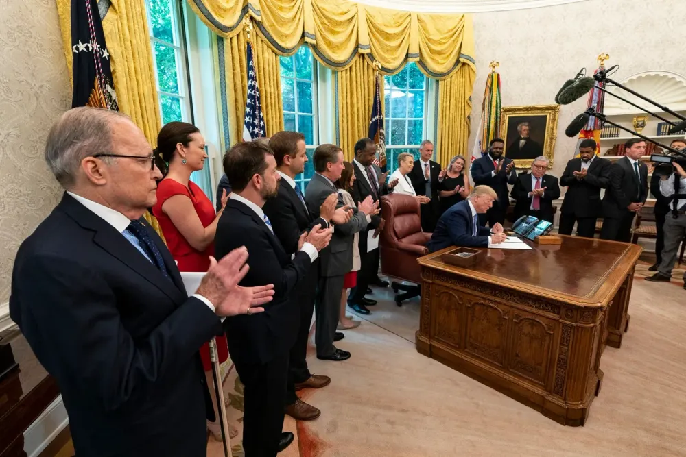 President Donald Trump signs a document at the Resolute Desk in the Oval Office while a group of people applauds.